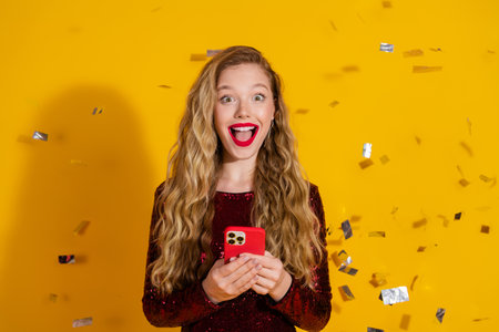 Stylish young woman in a red sequin dress holding a phone, smiling joyfully with confetti on a...