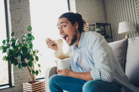 Young man enjoying a box of noodles in a cozy living room with natural lightingの写真素材