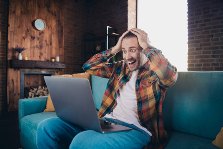 Excited young man in plaid shirt enjoying surprise on laptop while relaxing on sofa in stylish...
