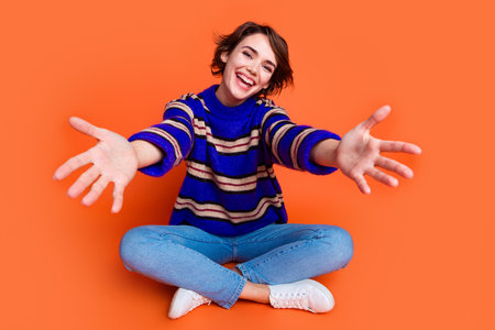 Photo portrait of young brown hair lady in blue striped cute sweater sitting floor welcome greetings to you isolated on orange color backgroundの写真素材