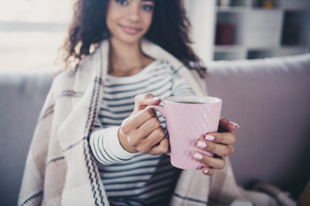 Cropped photo of attractive young woman sit couch wrapped blanket drinking cacao dressed striped outfit day light home interior living roomの写真素材