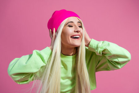 Young woman with blond hair in vibrant casual outfit and beanie smiling joyfully against a bright pink backgroundの写真素材