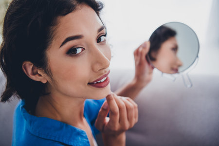 Beautiful young woman in casual blue shirt applying makeup using a small mirror while sitting comfortably in a brightly lit home interior.の写真素材