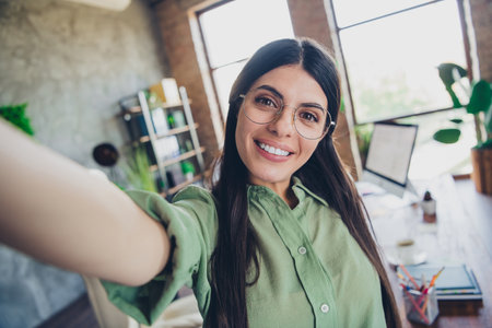 Confident young businesswoman with brunette hair taking selfie in modern office setting showing successの写真素材