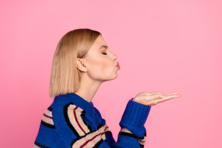 Stylish young woman in striped sweater blowing kiss with hand against pink background, expressing affection and joyの写真素材