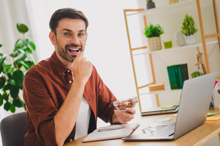 Smiling young man enjoying a light snack while working on a laptop in a cozy home office environment surrounded by plants and bookshelves.の写真素材