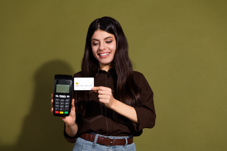Confident young woman holding a payment terminal and credit card against a trendy khaki backgroundの写真素材