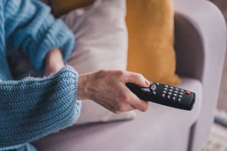 Elderly woman in blue sweater with remote control, relaxing indoors on sofaの写真素材