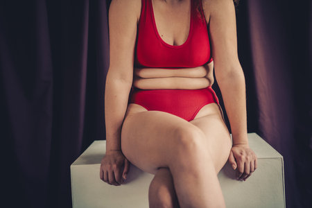 A young woman wearing natural and comfortable red lingerie, sitting confidently in a studio setting with dark purple curtainsの写真素材