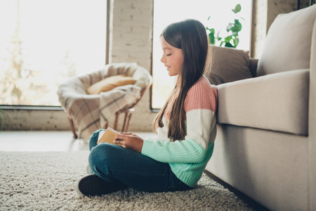 Young Girl Sitting on a Carpet in a Cozy Home Interior Reading a Book During a Relaxing Dayの写真素材