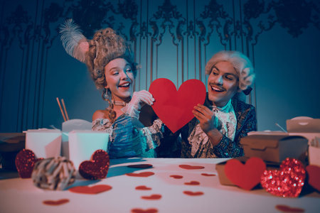 Elegant couple in baroque attire holding red heart in aristocratic setting with modern interior arrangementの写真素材