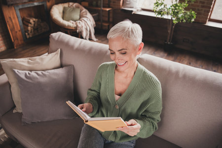Elderly woman enjoying a peaceful weekend at home with a charming smile, reading a book in her stylish green sweaterの写真素材