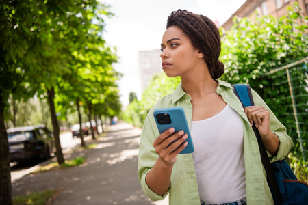 Young woman walking in sunny alley using smartphone and carrying backpack for travelの写真素材