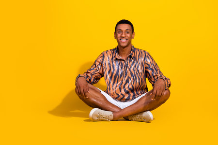 Young man in a trendy shirt sitting cross-legged against a vibrant yellow backgroundの写真素材