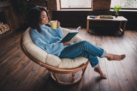 Young woman enjoying a relaxing day at home, reading a book and sipping coffee in a cozy living room chairの写真素材