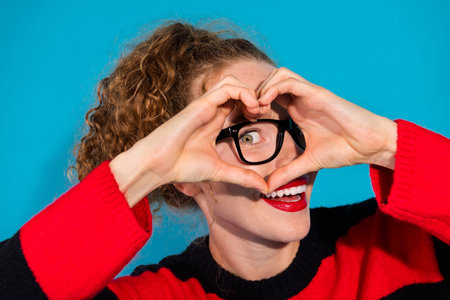 Young woman gestures heart shape with red striped sweater against blue backgroundの写真素材
