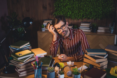 Young man studying late at night in a cozy home environment while surrounded by books and...