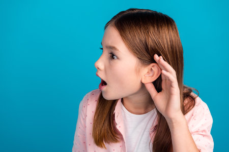 Young girl with curious expression listening attentively against blue background in a pink polka-dot shirtの写真素材