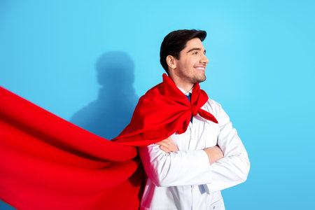 Confident young doctor wearing red cape in white coat standing against blue background symbolizing healthcare heroism and dedicationの写真素材