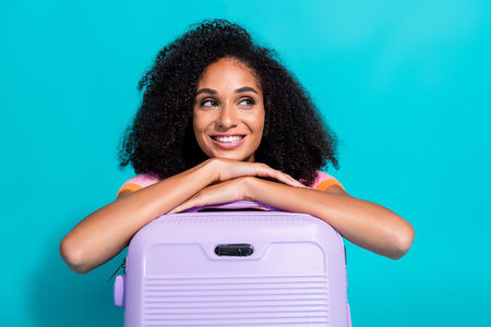 Happy woman with curly hair and stylish attire resting her arms on a violet suitcase against a vibrant teal backgroundの写真素材