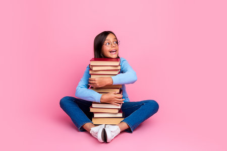Happy girl with glasses holding a pile of book, sitting on the floor against a vibrant pink background with a smileの写真素材