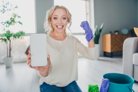 Cheerful woman celebrating success at home while holding a smartphone with empty screen and wearing casual clothes and glovesの写真素材