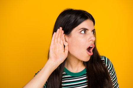 Surprised young woman cupping her ear against a vibrant yellow background, expressing curiosity and...