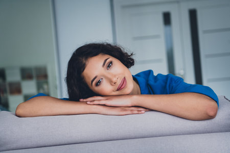 Young woman in casual blue shirt relaxing on couch with serene look in modern home interior during a...