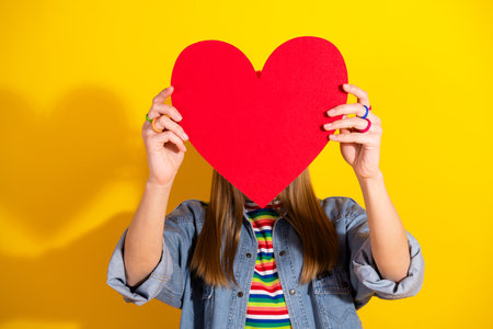 Young woman holding a red heart symbol against a vibrant yellow background, representing love and positive emotionsの写真素材
