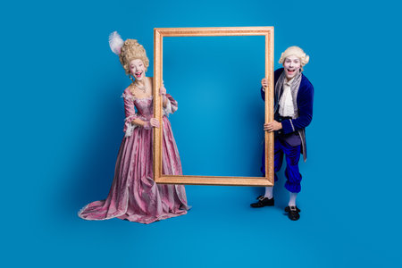 A joyful couple in elaborate baroque costumes poses with an empty frame against a vibrant blue...