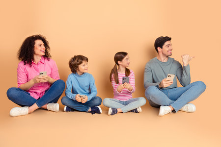 Happy family sitting on a beige background with smartphones showcasing laughter, bonding, and...