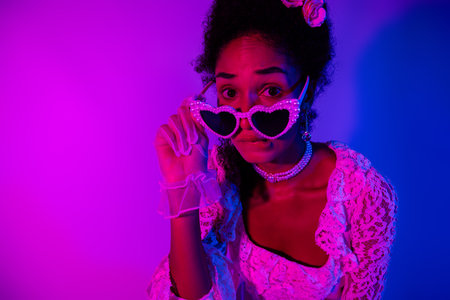 Young woman in elegant lace gown posing with heart-shaped sunglasses against a vibrant pink and...