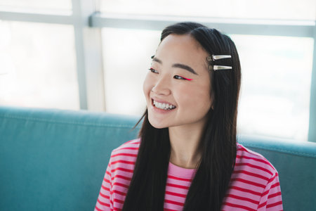 Young cheerful woman sitting indoors wearing pink striped shirt enjoying a casual relaxing day in a cozy and stylish living roomの写真素材