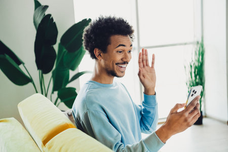Young man smiling during a video call while relaxing at home on a bright day with green plants in...