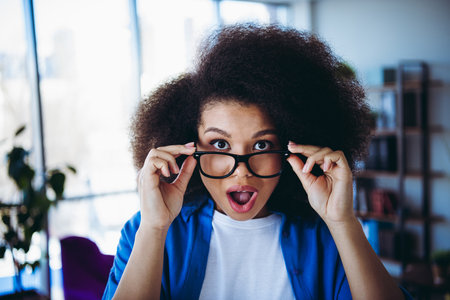 Young woman in a modern office environment looking surprised while adjusting her glassesの写真素材