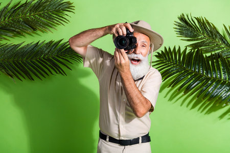 Photo of excited funny senior man photographer look take photo near palm leaves isolated green color backgroundの写真素材