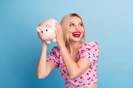 Young happy woman holding pink piggy bank, showcasing cheerful enthusiasm and financial planning against a bright blue backgroundの写真素材