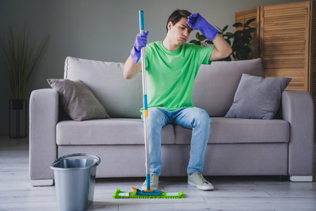 Young man in casual outfit mopping floor while sitting on a sofa in a cozy living room interior...