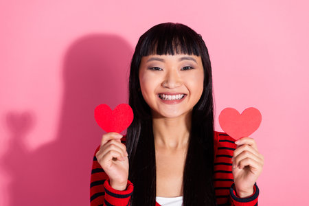 Smiling young woman holding two red hearts in hands against pink backdrop, expressing joy and loveの写真素材