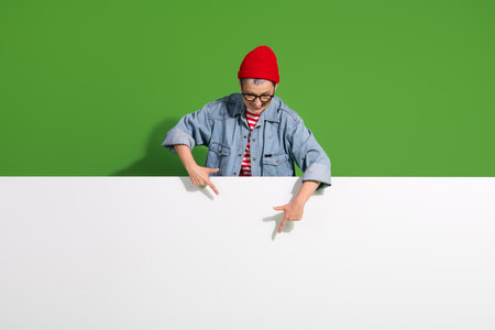 Confident senior woman wearing trendy denim and beanie pointing over blank space against green background for advertising ideasの写真素材