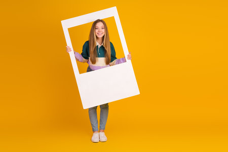 Young woman holding a photo frame smiling on a vibrant yellow background presenting cheerful style and casual charmの写真素材