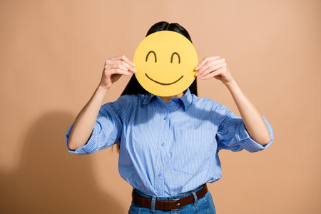 Young woman holding smiley face symbol showing positive emotions in casual attire against a beige backgroundの写真素材