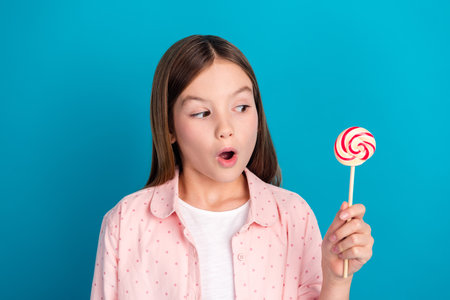Young girl with lollipop against a vivid blue background showcasing delight and curiosityの写真素材
