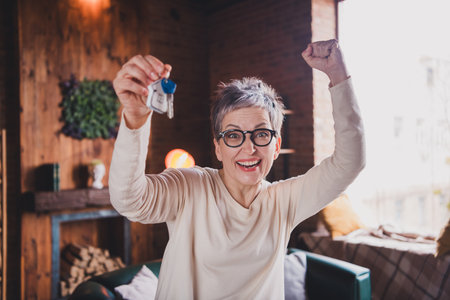 Elderly woman with grey hair celebrating at home, holding keys and smiling joyfully in a cozy indoor
