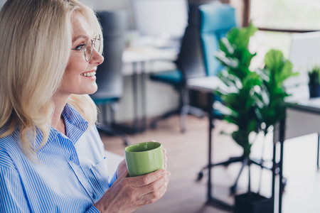 Mature businesswoman enjoying a coffee break in a modern office setting with natural daylightの写真素材