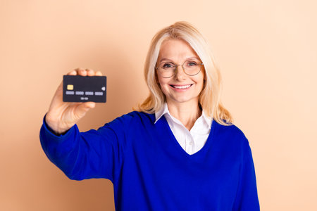 Confident mature businesswoman holding a credit card with a radiant smile on beige backgroundの写真素材