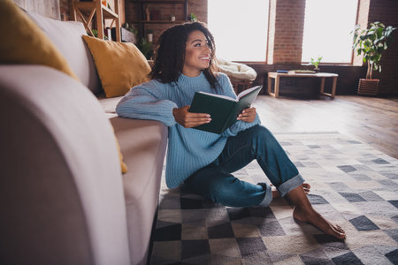 Young woman in cozy sweater sitting on carpet, smiling while reading a book in stylish living room with natural daylightの写真素材