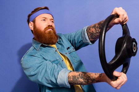 Stylish bearded man holding a steering wheel, wearing a denim jacket with tattoos and a headband