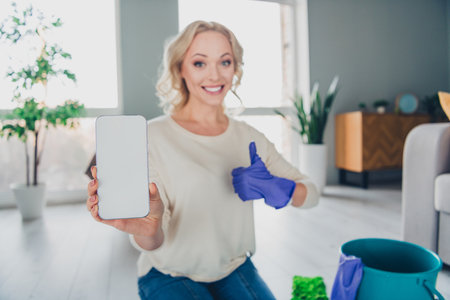 Young Woman in a Bright Living Room Showing a Smartphone and Expressing Positivity With a Confident...