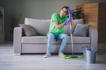 Young man cleaning living room with mop, expressing exhaustion, wearing casual attire, sitting on sofaの写真素材